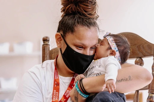 A woman wearing a face mask holding a baby, seeking rescue from homelessness with the support of Open Door Mission.